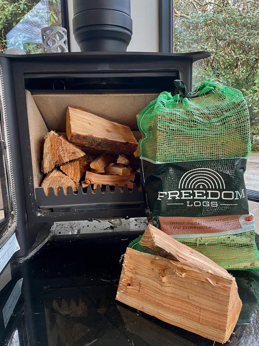 A set of kiln dried logs in a net bag, with some logs placed inside a wood stove, situated in a room with a glass window looking out to a green outdoor scene.